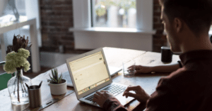 Man sitting at a desk working on a laptop with another person to his right.