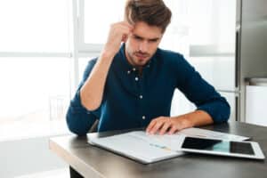 Photography of concentrated young man sitting at home and analyzing his finances. Looking at documents and touching his head.