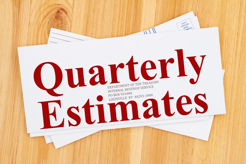 Quarterly Estimates in Red letters on a white envelope addressed to the US Treasury in Kentucky laying on a wood table. Quarterly tax payments are required if you are self-employed.