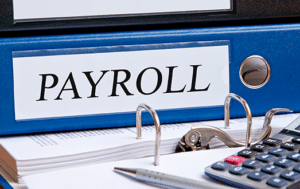 Text payroll in uppercase black letters on blue binder with pen and electronic calculator placed alongside. 2025 Payroll Changes - Brown, Chism & Thompson